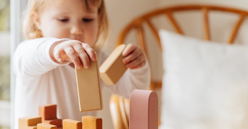 Cute little girl in white casual clothes standing near table and playing with wooden blocks while spending time at home