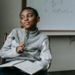 Concentrated African American boy in casual outfit sitting on chair with pencil and copybook and thinking how to do task